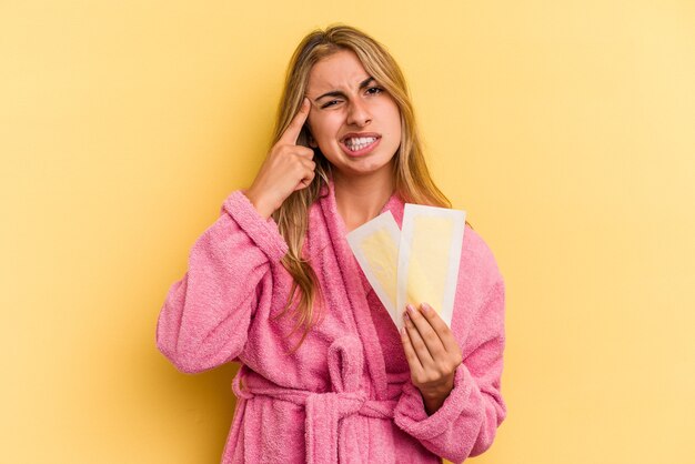 Jeune femme blonde caucasienne portant un peignoir tenant des bandes dépilatoires isolées sur fond jaune montrant un geste de déception avec l'index.