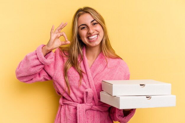 Jeune femme blonde caucasienne portant un peignoir tenant des pizzas isolées sur fond jaune joyeux et confiant montrant un geste ok.