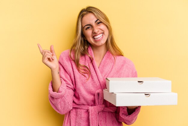 Jeune femme blonde caucasienne portant un peignoir tenant des pizzas isolées sur fond jaune souriant et pointant de côté, montrant quelque chose dans un espace vide.