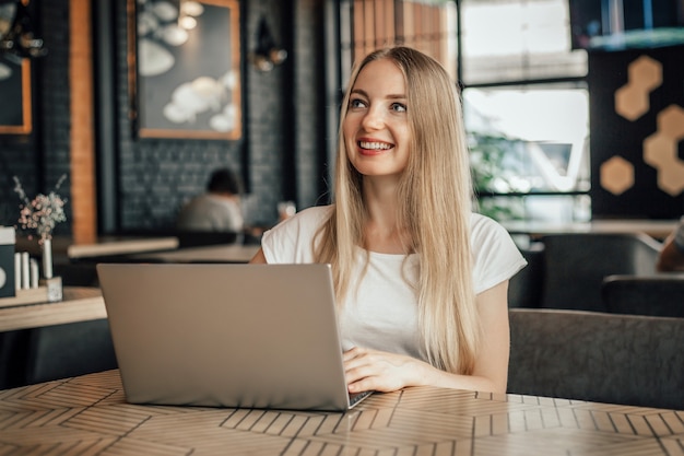 Jeune femme blonde caucasienne souriante et assise avec un ordinateur portable à une table dans un café détourner le regard et travailler