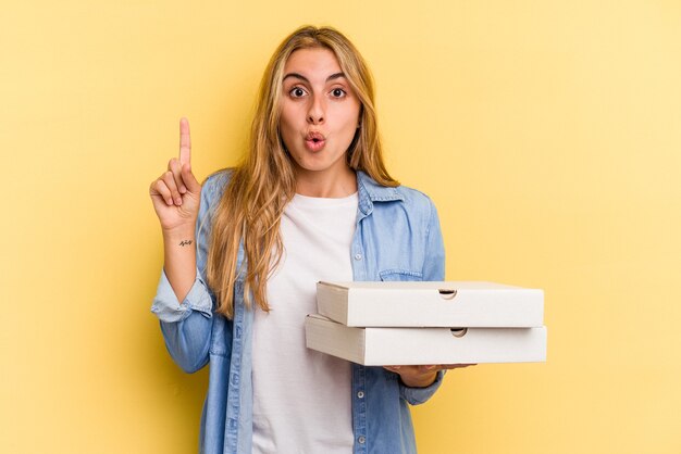 Jeune femme blonde caucasienne tenant des pizzas isolées sur fond jaune ayant une bonne idée, concept de créativité.