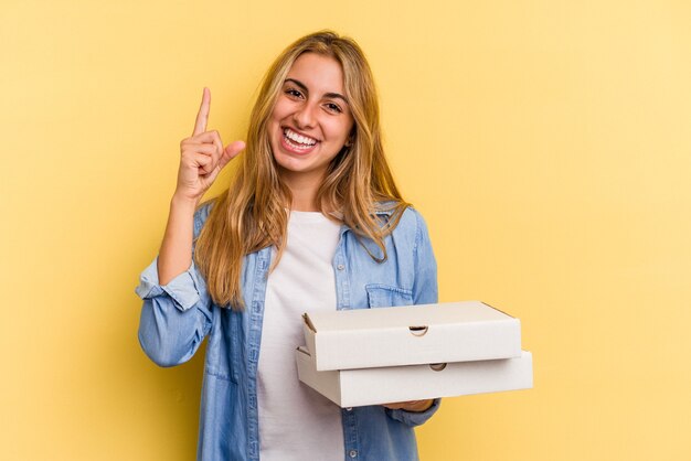 Jeune femme blonde caucasienne tenant des pizzas isolées sur fond jaune montrant le numéro un avec le doigt.