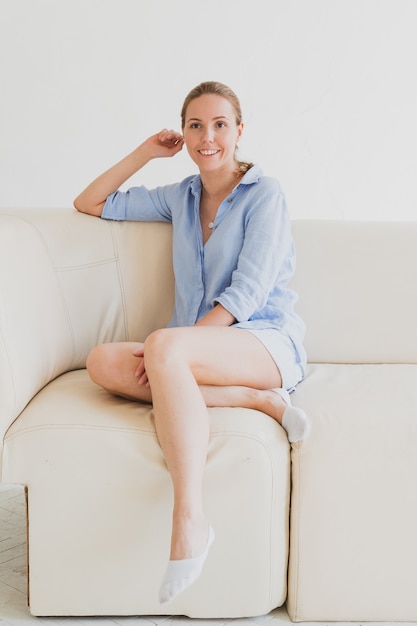 Jeune femme blonde en chemise bleue est assise sur un canapé dans une pièce lumineuse et rit