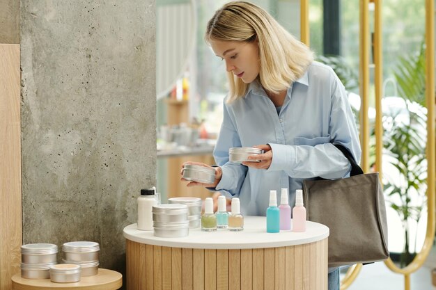 Jeune femme blonde choisissant une crème pour le corps ou un gommage dans un pot en étain au supermarché