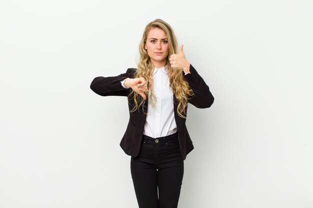 Jeune femme blonde, confuse, désemparée et incertaine, soupesant le bien et le mal entre différentes options ou choix contre un mur blanc