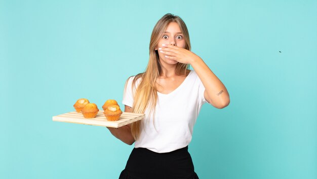 Jeune femme blonde couvrant la bouche avec les mains avec un choqué et tenant un muffins troy