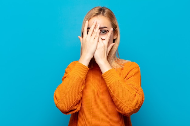 Jeune femme blonde couvrant le visage avec les mains, furtivement entre les doigts avec une expression surprise et regardant sur le côté