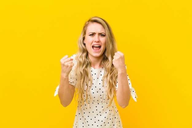 Jeune femme blonde criant agressivement avec une expression de colère ou avec les poings serrés célébrant le succès contre le mur jaune