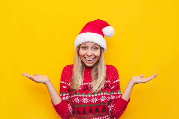 Une jeune femme blonde dans un chandail chaud de cerf rouge et un chapeau de père Noël écarte les mains avec le doute