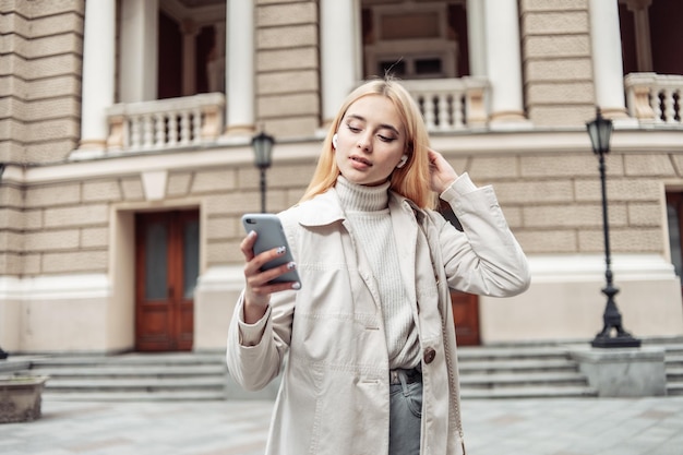 Photo jeune femme blonde dans les écouteurs à l'aide de smartphone en ville