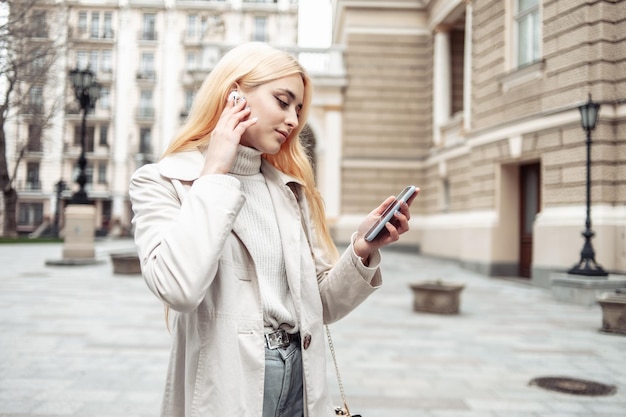 Jeune femme blonde dans les écouteurs à l'aide de smartphone en ville