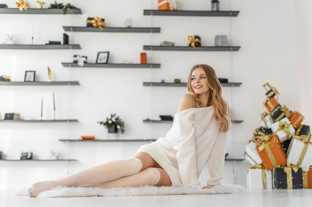 jeune femme blonde dans un long pull confortable est assise sur le sol en profitant du confort de sa maison