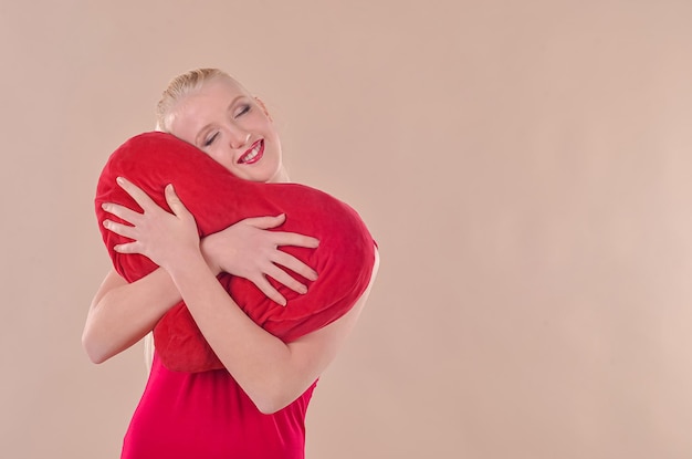 La jeune femme blonde dans une robe rouge tient un coeur rouge dans des ses mains sur un fond beige