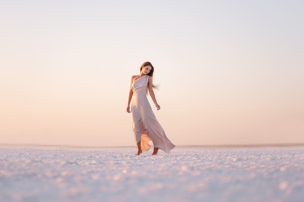 Jeune femme blonde dans une soirée rose pastel aéré, robe poudrée se tient pieds nus sur du sel cristallisé blanc. Excursion minière de sel, marche sur l'eau au coucher du soleil