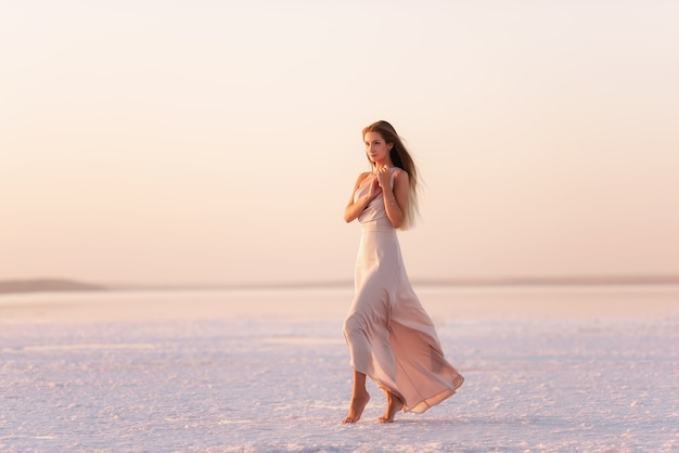 Photo jeune femme blonde dans une soirée rose pastel aéré, robe poudrée se tient pieds nus sur du sel cristallisé blanc. excursion minière de sel, marche sur l'eau au coucher du soleil