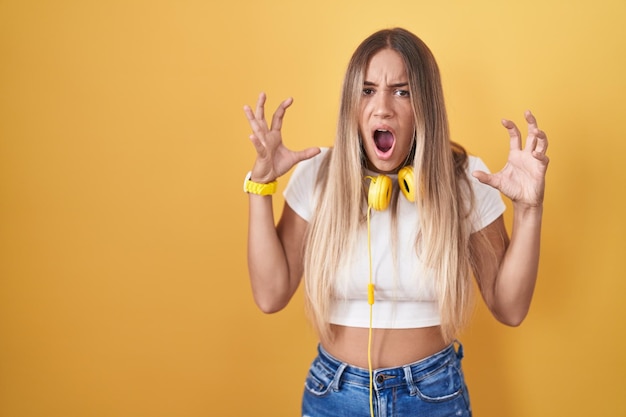 Jeune femme blonde debout sur fond jaune portant des écouteurs fous et fous criant et hurlant avec une expression agressive et les bras levés concept de frustration