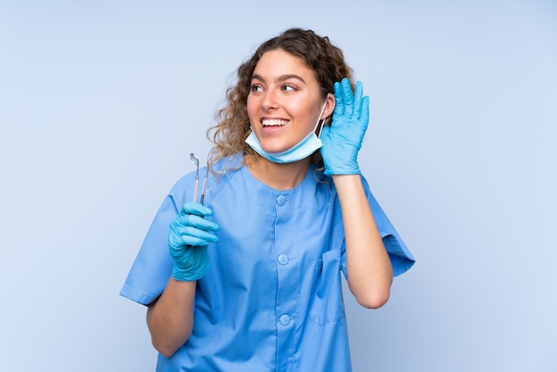 Jeune femme blonde dentiste tenant des outils sur le mur bleu en écoutant quelque chose