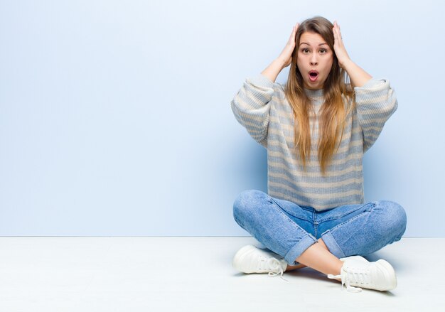 Jeune femme blonde à la désagréablement choquée, effrayée ou inquiète, la bouche grande ouverte et couvrant les deux oreilles avec les mains assis sur le sol