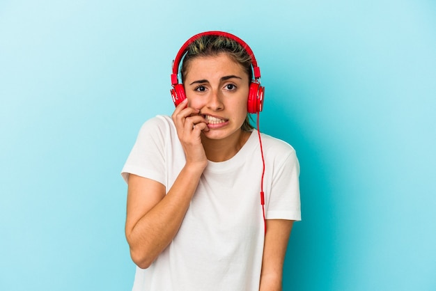 Jeune femme blonde écoutant de la musique sur un casque isolé sur un mur bleu se ronger les ongles, nerveux et très anxieux