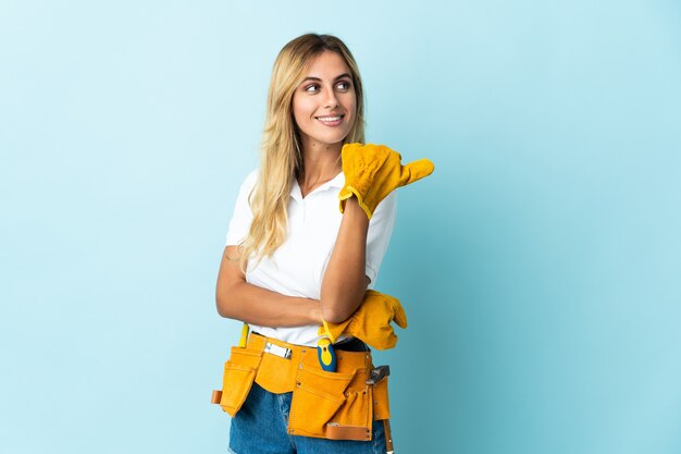 Jeune femme blonde électricien uruguayen isolé sur un mur bleu pointant vers le côté pour présenter un produit