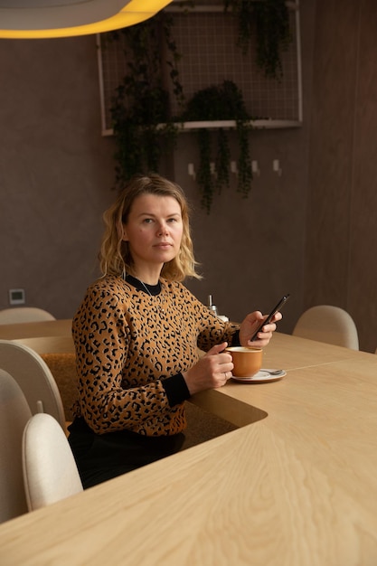 Une jeune femme blonde est assise dans un café dans des tons beiges buvant du café défilant sur le téléphone et souriant