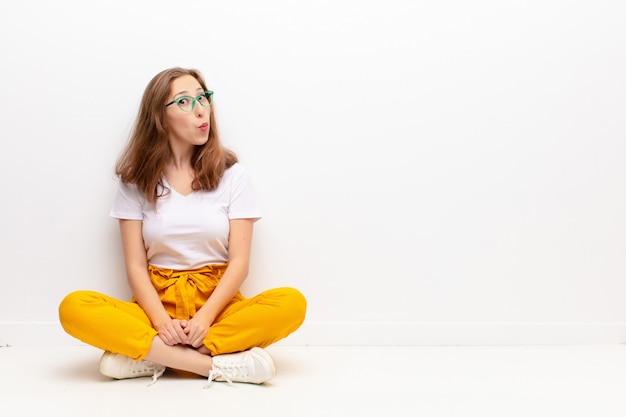 Photo jeune femme blonde avec une expression maladroite, folle et surprise, les joues gonflées, se sentant bourré, gras et plein de nourriture assis sur le sol