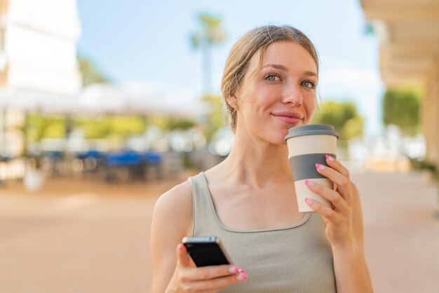 Jeune femme blonde à l'extérieur à l'aide de téléphone portable et tenant un café