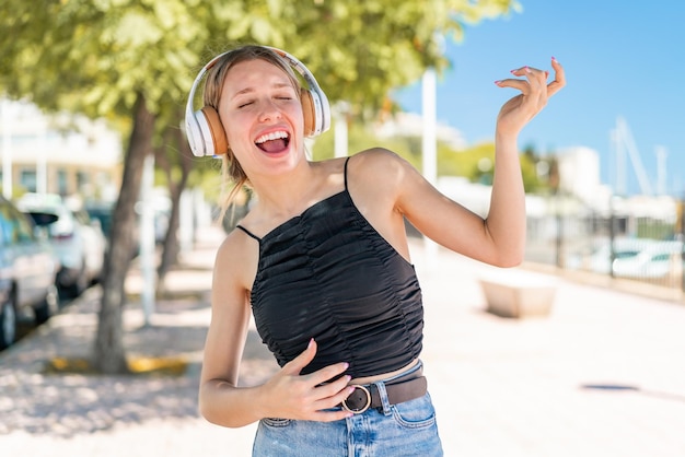 Jeune femme blonde à l'extérieur écoutant de la musique et faisant un geste de guitare