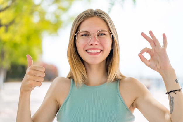 Photo jeune femme blonde à l'extérieur avec des lunettes et faisant signe ok