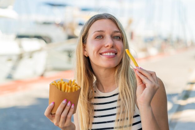 Jeune femme blonde à l'extérieur prenant des frites frites avec une expression heureuse