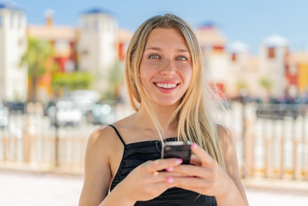 Jeune femme blonde à l'extérieur regardant la caméra et souriant tout en utilisant le mobile