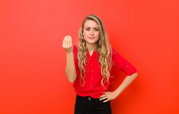 Jeune femme blonde faisant un geste de capice ou d'argent, vous disant de payer vos dettes! contre le mur rouge