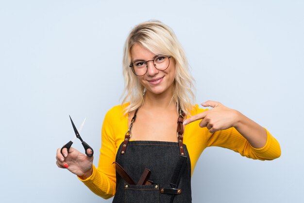 Jeune femme blonde sur fond isolé avec une robe de coiffeur ou un coiffeur et en le pointant