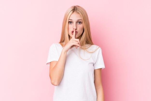 Jeune femme blonde sur fond rose gardant un secret ou demandant le silence.