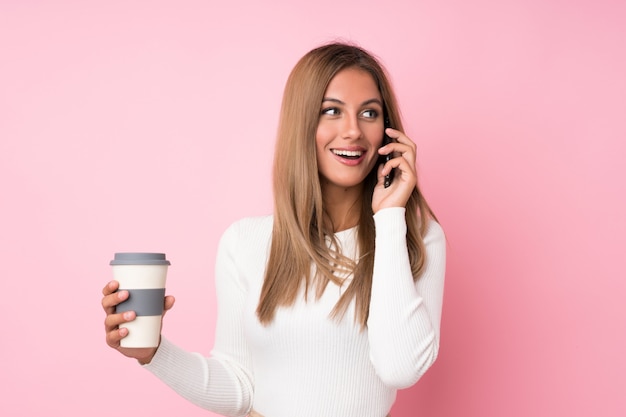 Jeune femme blonde sur fond rose isolé tenant un café à emporter et un téléphone portable