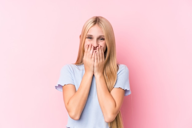 Jeune femme blonde sur fond rose riant de quelque chose, couvrant la bouche avec les mains.