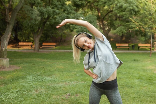 Une jeune femme blonde en forme se réchauffe avant de s'entraîner et écoute de la musique sur des écouteurs dans le parc. Aérobic