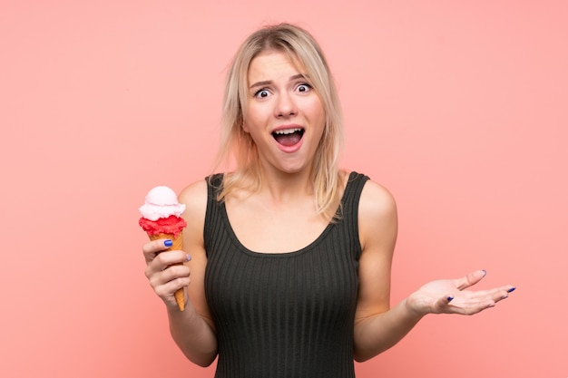 Jeune femme blonde avec une glace au cornet sur un mur rose isolé avec une expression faciale choquée