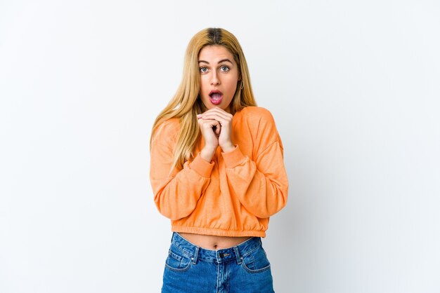 Jeune femme blonde isolée sur blanc priant pour la chance, étonné et ouvrant la bouche à l'avant.