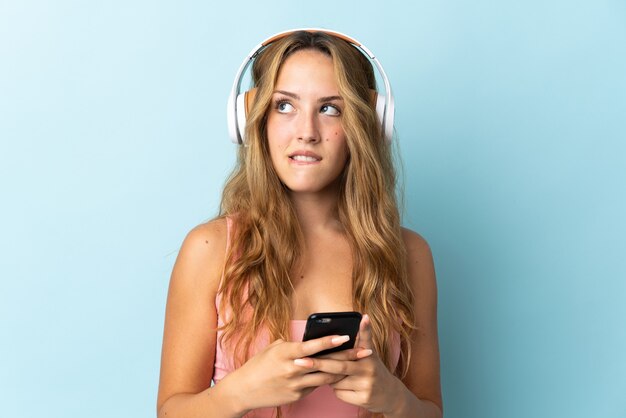 Jeune femme blonde isolée sur bleu écoute de la musique avec un mobile et de la pensée