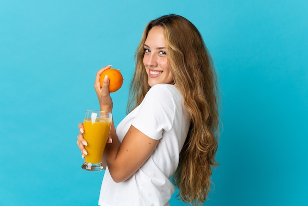 Jeune femme blonde isolée sur fond bleu tenant une orange et un jus d'orange