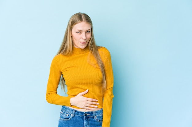 Jeune femme blonde isolée sur fond bleu touche le ventre, sourit doucement, concept de manger et de satisfaction.