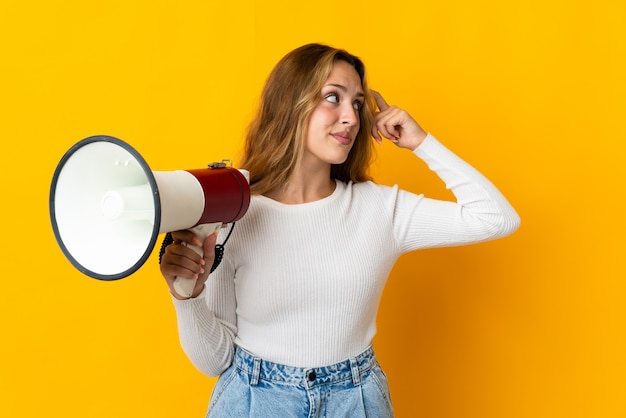 Jeune femme blonde isolée sur fond jaune tenant un mégaphone et ayant des doutes