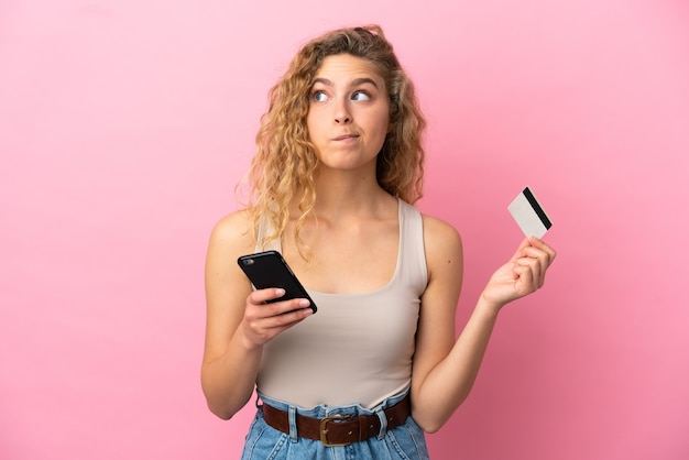 Jeune femme blonde isolée sur fond rose achetant avec le mobile avec une carte de crédit en pensant
