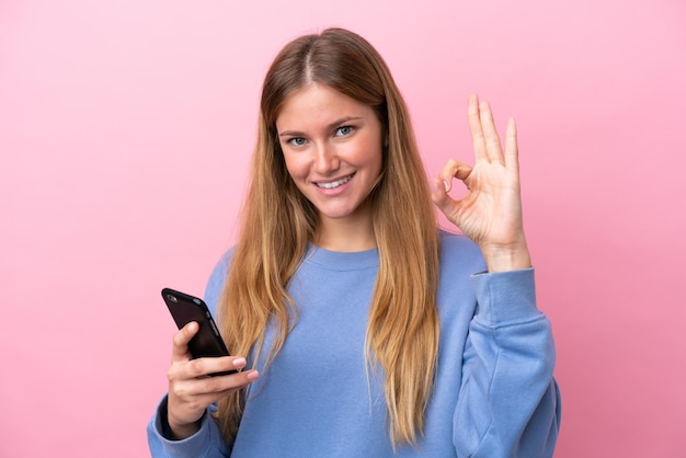 Jeune femme blonde isolée sur fond rose à l'aide d'un téléphone portable et faisant signe OK