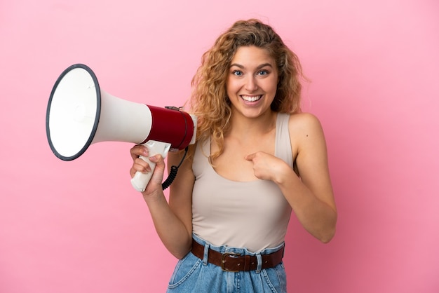 Jeune femme blonde isolée sur fond rose tenant un mégaphone et avec une expression faciale surprise