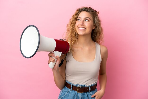 Jeune femme blonde isolée sur fond rose tenant un mégaphone et levant les yeux en souriant