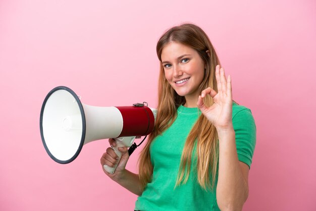 Jeune femme blonde isolée sur fond rose tenant un mégaphone et montrant le signe ok avec les doigts