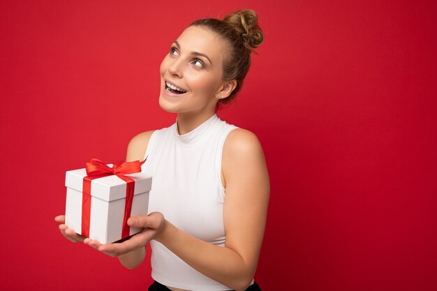 jeune femme blonde isolée sur fond rouge mur portant haut blanc tenant une boîte-cadeau et regardant sur le côté et de la pensée. Espace libre