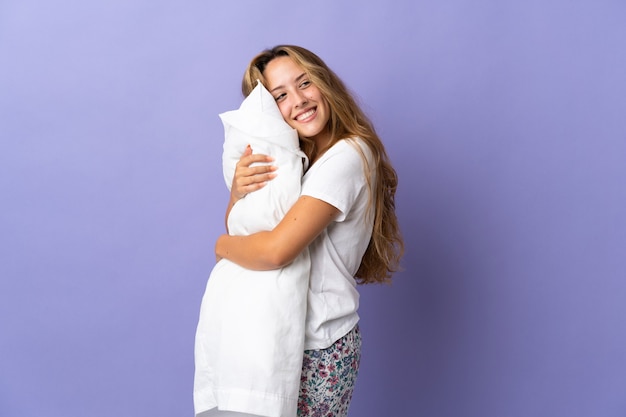 Jeune femme blonde isolée sur fond violet en pyjama et tenant un oreiller avec une expression heureuse